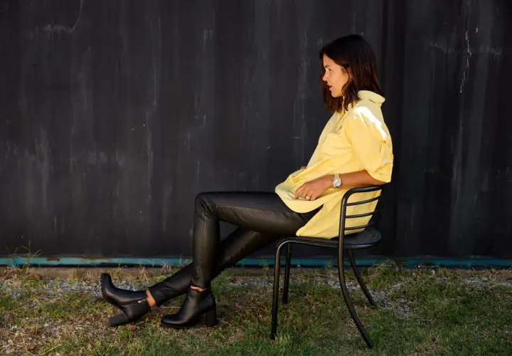 Une femme assise porte un pantalon avec bottines homme, une chemise jaune vif et une montre.