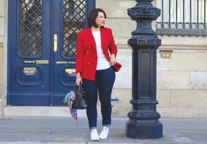 Une femme élégante montre comment porter un blazer rouge femme avec un jean et des baskets. Elle tient un sac noir orné d'un foulard coloré.