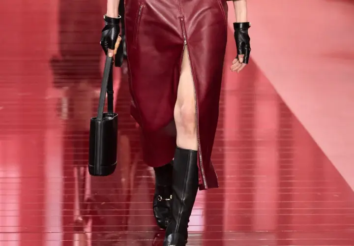 Mannequin portant une jupe courte en cuir bordeaux fendue et des bottines noires. Sac cylindrique noir et gants en cuir.