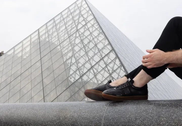 Un homme porte une tenue avec basket noir, assis devant la pyramide du Louvre.