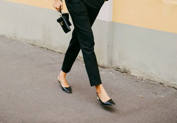 Une femme élégante marche dans la rue, portant un pantalon noir et des chaussures ballerines noires. Elle tient un petit sac noir.