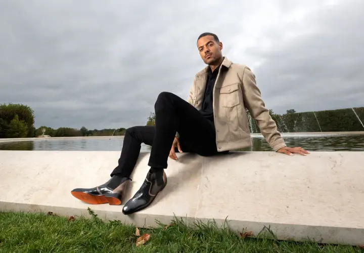 Un homme élégant montre comment porter des chelsea boots homme avec un jean noir et une veste beige, près d'une fontaine.
