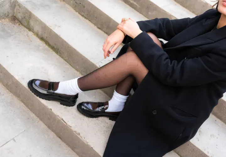 Une personne assise sur des marches de béton porte un manteau noir, des collants noirs et des chaussettes blanches avec des chaussures de type chaussette mocassin vernies.