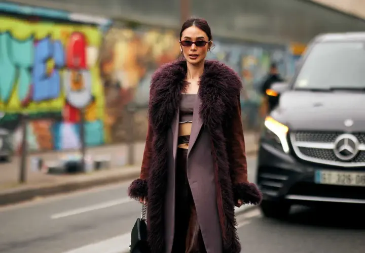 Une femme porte un manteau long en fausse fourrure prune, un exemple de types de manteaux femme tendance. Elle tient un sac noir.