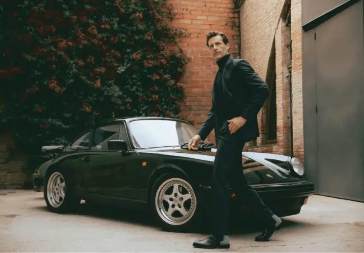 Un homme élégant en costume et bottines noires pose près d'une Porsche verte.
