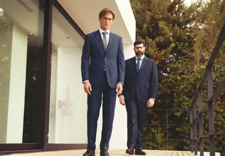 Deux hommes en costume sans chaussette, l'un grand et mince, l'autre plus trapu, posent sur une terrasse en bois devant une maison moderne.