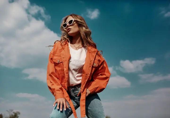 Une jeune femme porte une tenue mi saison femme stylée : veste orange, t-shirt blanc et jean. Elle est dehors, sous un ciel bleu nuageux.