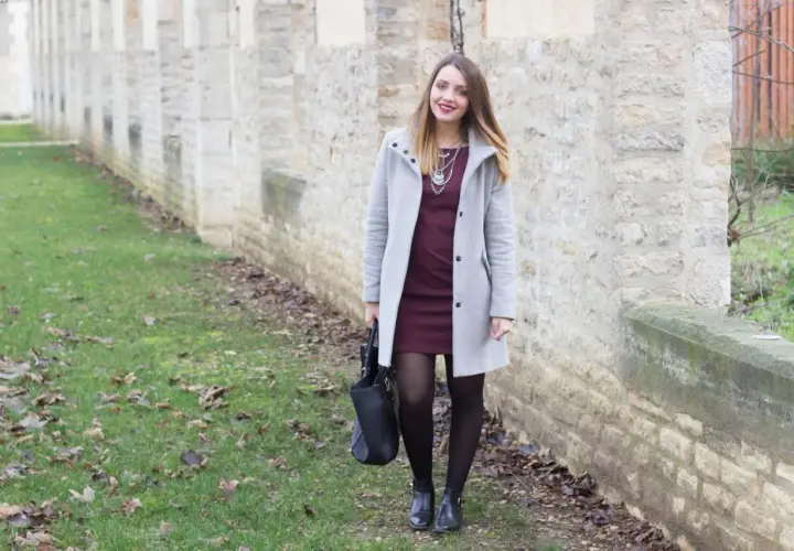Une femme porte une robe bordeaux et un manteau gris clair. L'association couleur bordeaux vêtements est élégante.