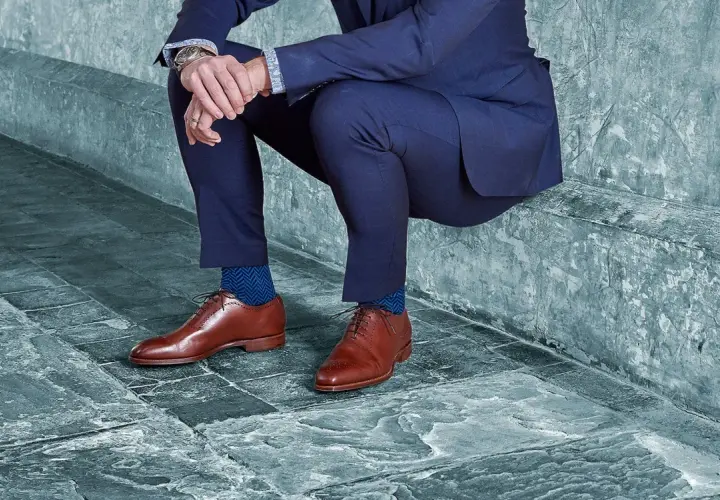 Un homme en costume bleu, avec des chaussures noires et des chaussettes bleues à chevrons, est assis sur un banc en pierre.