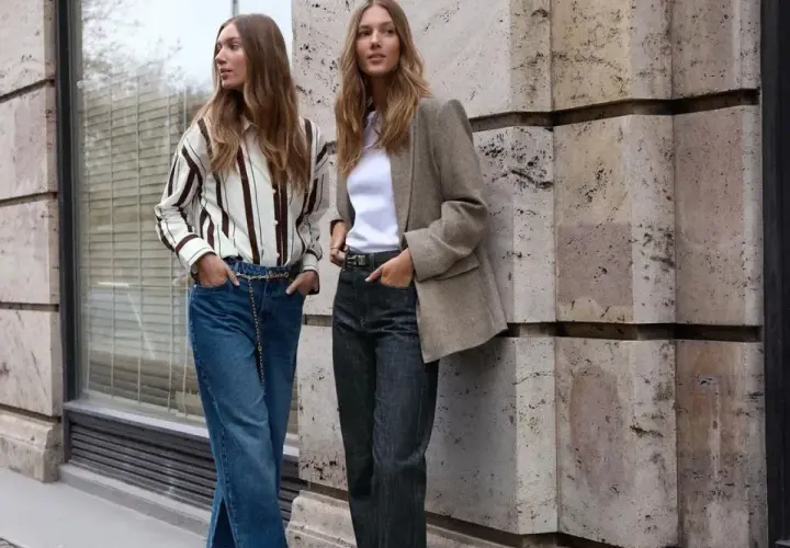 Deux femmes posent devant un mur en pierre. L'une porte un jean tapered femme, une chemise rayée et une ceinture chaîne. L'autre, un blazer et un jean.
