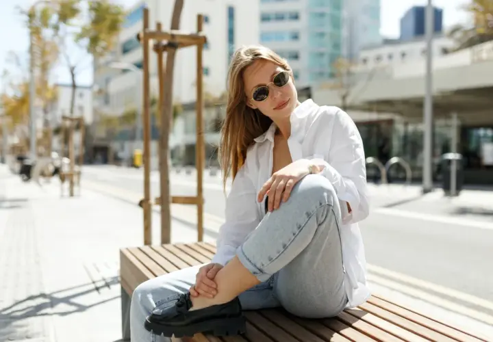 Une femme assise sur un banc, portant un chemisier blanc, un jean décontracté et des mocassins noirs. Son outfit simple et chic est parfait pour une journée en ville.