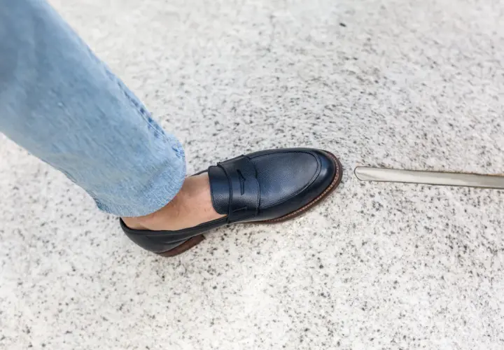 Un pied en jean bleu porte un mocassin bleu marine. La semelle extérieure de la chaussure, signée par un cordonnier, est visible.
