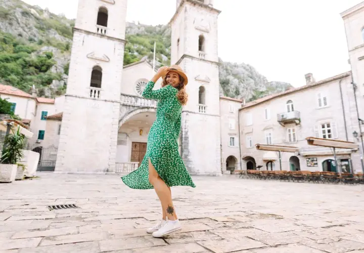 Une femme porte une robe verte fleurie et des baskets blanches, un chapeau de paille sur la tête, dans une place pavée devant une église.