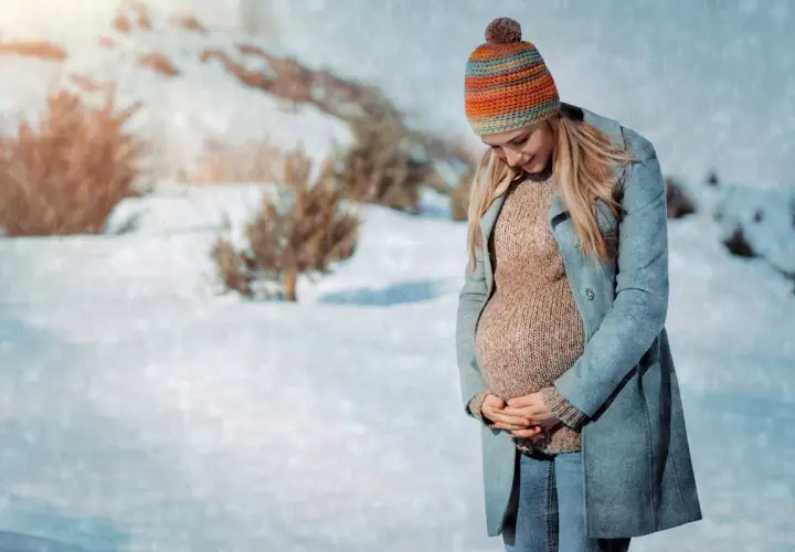 Une femme enceinte, bien emmitouflée dans un manteau et un bonnet coloré, pose dans un paysage enneigé. Elle montre comment s'habiller enceinte en hiver avec style et confort.