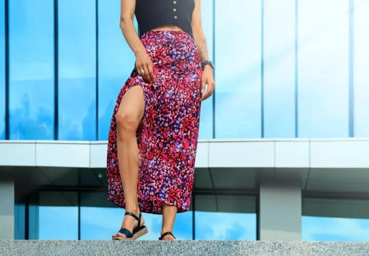 Une femme en jupe longue à fleurs et escarpins bleus, posant devant un bâtiment moderne aux reflets bleus.