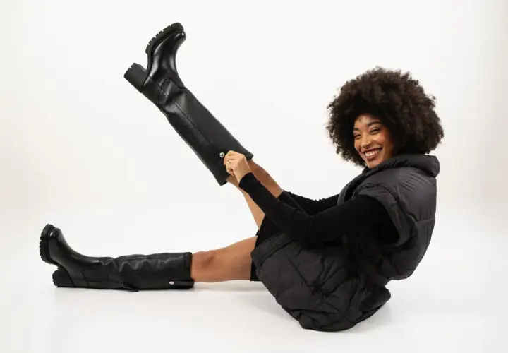 Une femme souriante pose avec une tenue noire, mettant en valeur ses bottes à talons hautes et épaisses.
