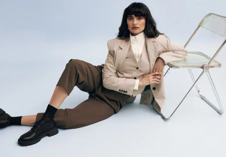 Femme en tenue professionnelle, blazer beige, pantalon marron, et chaussure entretien d'embauche femme noire. Assise près d'une chaise transparente.