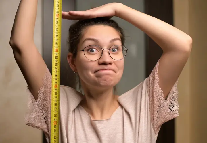 Une femme trapue, lunettes rondes, mesure sa taille avec un mètre ruban jaune. Elle a une expression amusée.