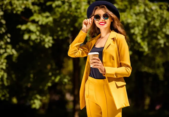 Une femme en costume jaune vif, chapeau et lunettes de soleil, tenant un café. Parfait pour savoir comment s'habiller quand il fait 14 degrés.