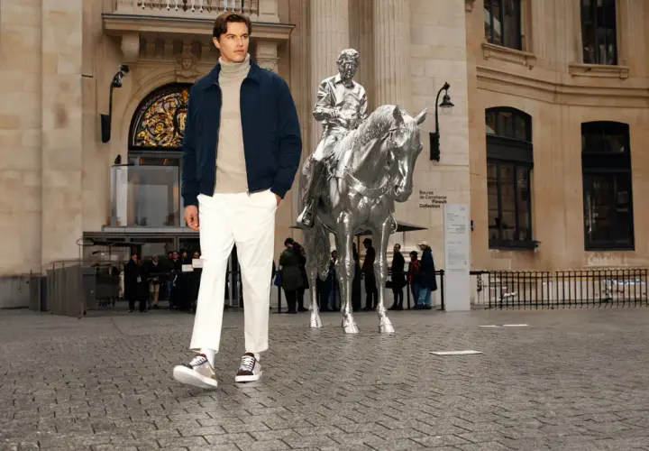 Un homme porte une chaussure avec un chino blanc, un pull à col roulé beige et une veste bleu marine devant une sculpture de cheval argenté.
