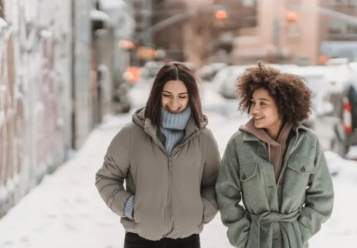 Deux amies discutent en marchant dans la neige. Une idée outfit hiver parfaite : doudoune beige et manteau vert ceinturé.