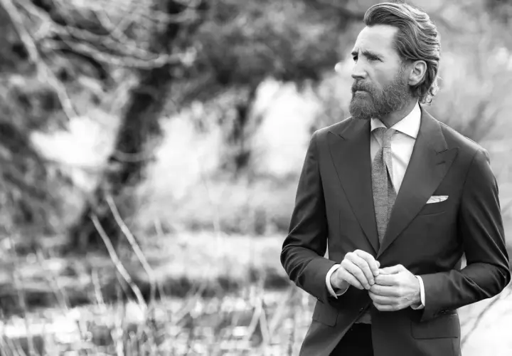 Un homme barbu en costume sombre, chemise blanche et cravate grise, ajuste sa veste. Sa tenue avec short blanc homme est élégante.