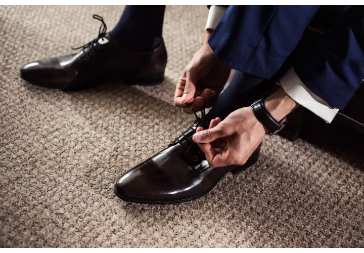 Un homme en costume bleu marine attache ses chaussures vernies marron. Quel chaussure avec un costume pour un look impeccable.