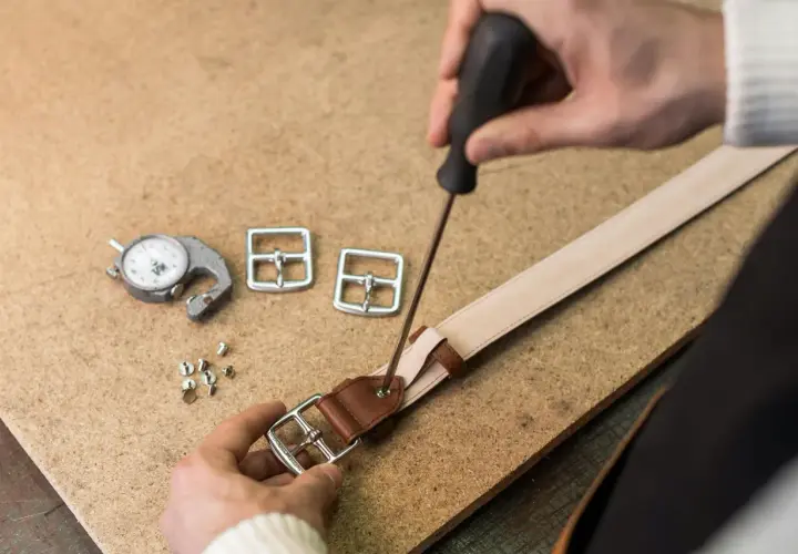 Artisan travaillant sur une ceinture en cuir, utilisant un tournevis pour fixer une boucle. Outils de mesure et boucles supplémentaires à proximité.