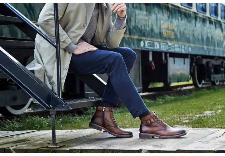 Un homme élégant, assis près d'un train, porte des bottines marron. Une inspiration pour savoir quelles chaussures porter avec un jean homme.