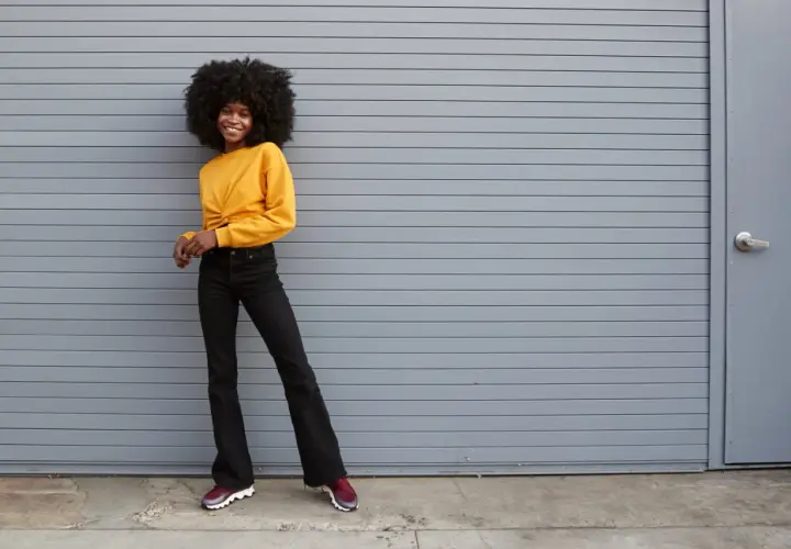 Une jeune femme souriante pose devant un mur gris, portant un haut jaune et un jean flare. Quelles chaussures avec jean flare ? Des baskets bordeaux complètent ce look décontracté.