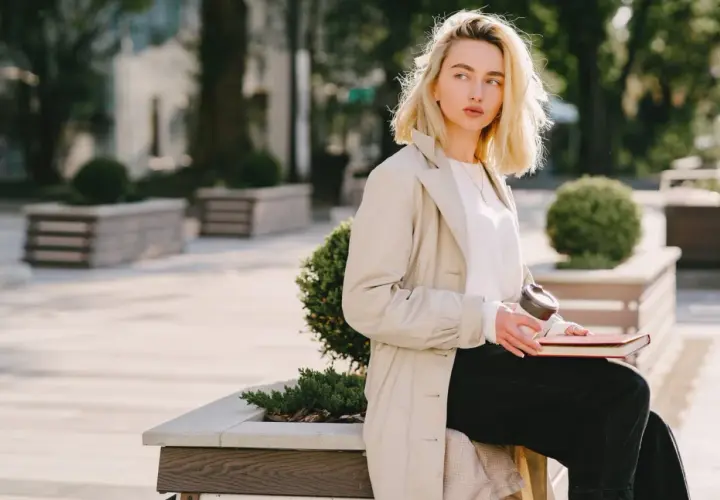 Une femme blonde, assise dehors avec un livre et un café, porte un trench beige et un haut blanc. Parfait pour savoir comment s'habiller quand il fait 15 degrés.