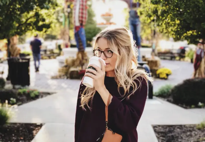 Une femme blonde avec des lunettes boit un café. Elle porte un pull bordeaux. Elle se demande comment dois je m habiller demain.