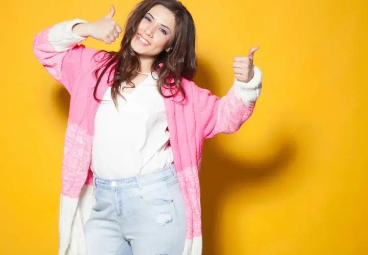 Une femme souriante, petite et ronde, montre comment s'habiller avec un gilet rose et un jean. Elle fait un signe de pouce levé.