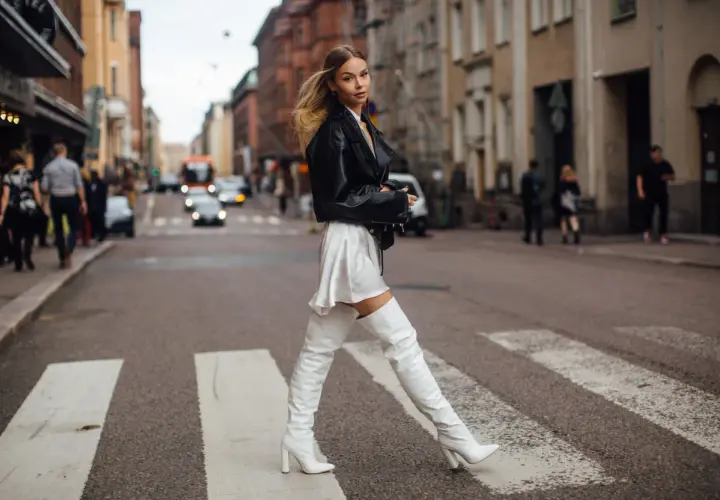 Une femme traverse la rue dans une tenue audacieuse : veste en cuir noire, mini-jupe blanche et cuissardes plates blanches.
