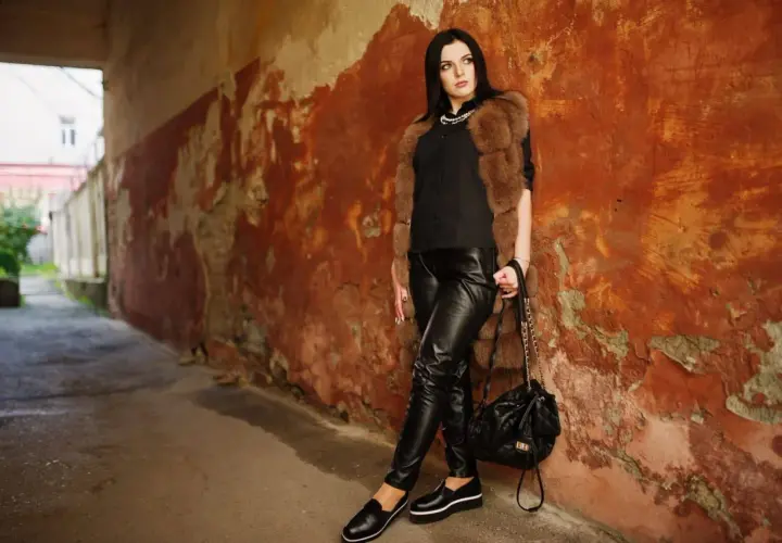 Une femme pose contre un mur décrépit, vêtue d'un pantalon en cuir noir. Apprenez comment nettoyer un pantalon en cuir pour qu'il reste aussi beau.