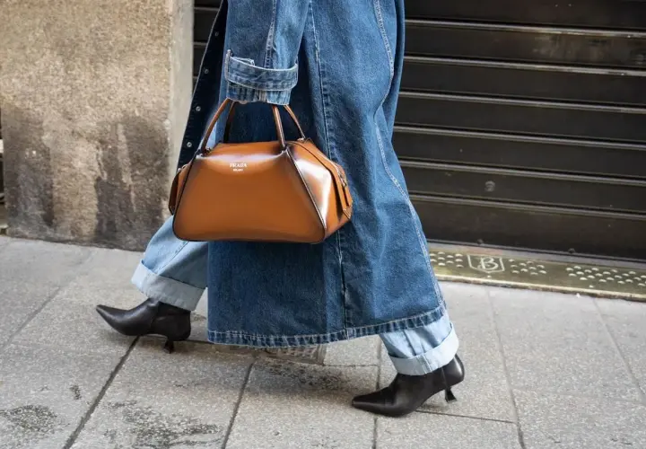 Une femme élégante marche dans la rue, portant un jean retroussé, une veste en jean et un sac à main marron. Le style "comment retrousser son jean femme" est parfait pour cette tenue.