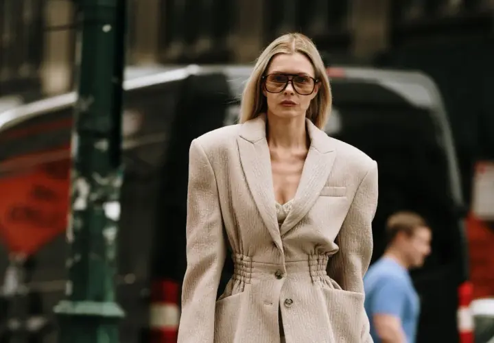 Une femme porte une tenue avec un blazer blanc chic, des lunettes de soleil oversize et un top en dentelle. Elle déambule dans la rue.
