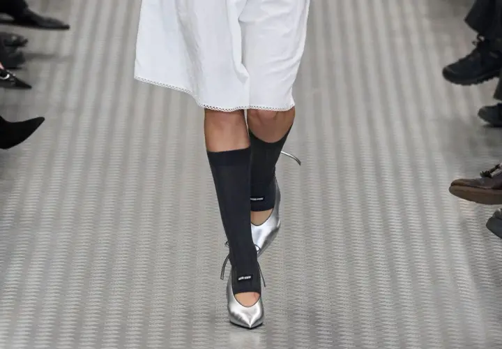 Une mannequin défile avec une tenue blanche et des chaussettes hautes grises, rehaussée par des chaussures argentées pointues.