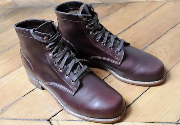 Bottes marron en cuir gras, lacets gris, sur un parquet en bois.