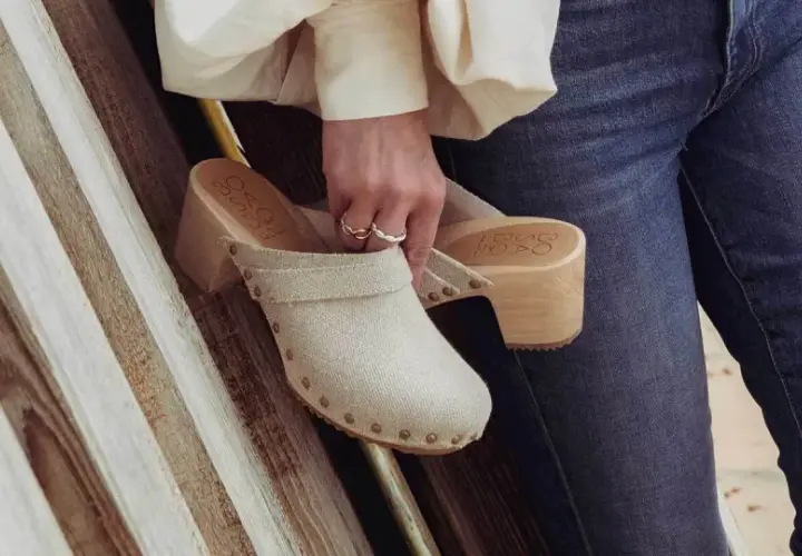 Une femme tient un sabot femme en toile beige, orné de clous, avec un talon en bois. Elle porte un jean bleu.