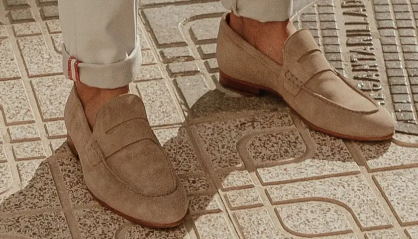 Un homme porte un mocassin à la mode en daim beige, avec un pantalon retroussé, sur un trottoir en dalles.