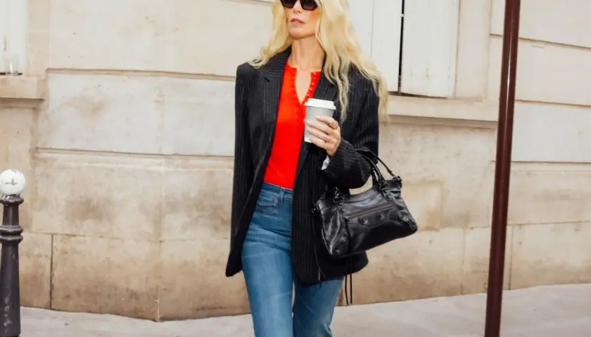 Femme blonde portant un jean évasé et un blazer rayé, avec un haut rouge vif et un sac à main noir.