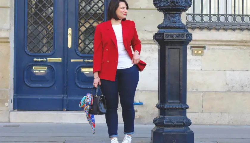 Une femme élégante montre comment porter un blazer rouge femme avec un jean et des baskets. Elle tient un sac noir orné d'un foulard coloré.
