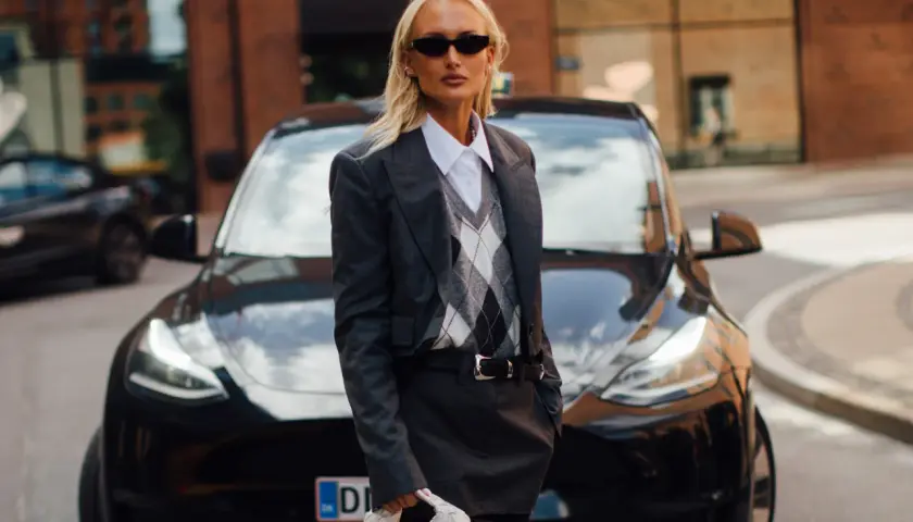 Une femme stylée devant une Tesla noire, portant un ensemble blazer et jupe gris. Un look parfait pour savoir quel blazer pour quelle morphologie.
