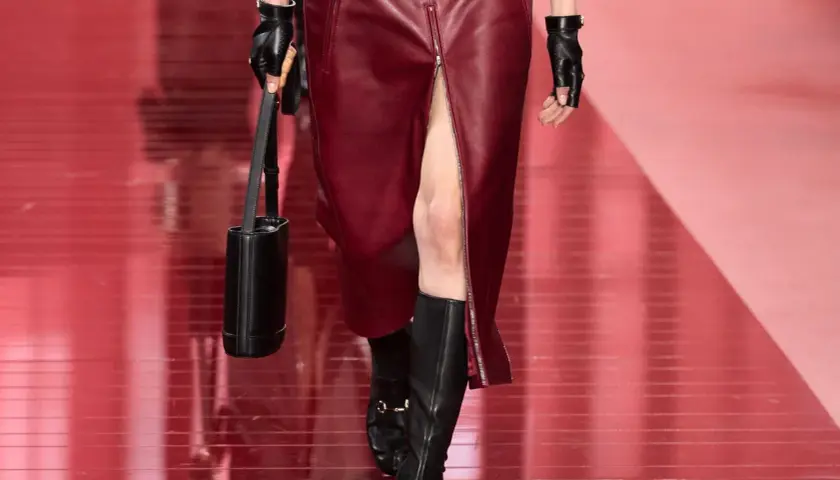 Mannequin portant une jupe courte en cuir bordeaux fendue et des bottines noires. Sac cylindrique noir et gants en cuir.