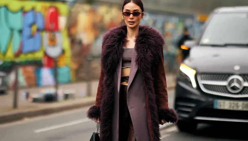 Une femme porte un manteau long en fausse fourrure prune, un exemple de types de manteaux femme tendance. Elle tient un sac noir.