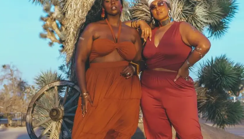 Deux femmes aux courbes généreuses posent dans le désert. L'une porte une robe longue orange, l'autre un ensemble pantalon et top rouge. Des tenues parfaites pour savoir comment s'habiller quand on a des grosses cuisses.