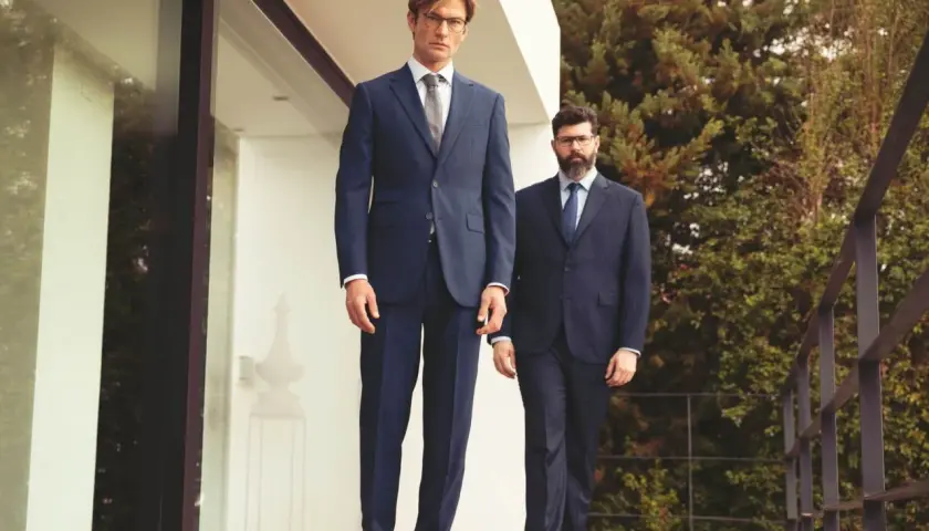 Deux hommes en costume sans chaussette, l'un grand et mince, l'autre plus trapu, posent sur une terrasse en bois devant une maison moderne.