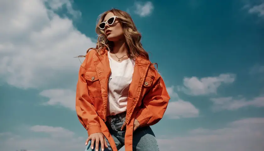 Une jeune femme porte une tenue mi saison femme stylée : veste orange, t-shirt blanc et jean. Elle est dehors, sous un ciel bleu nuageux.