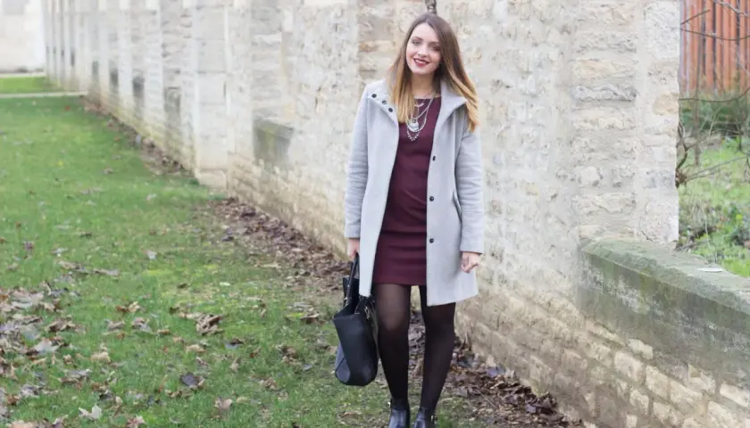 Une femme porte une robe bordeaux et un manteau gris clair. L'association couleur bordeaux vêtements est élégante.